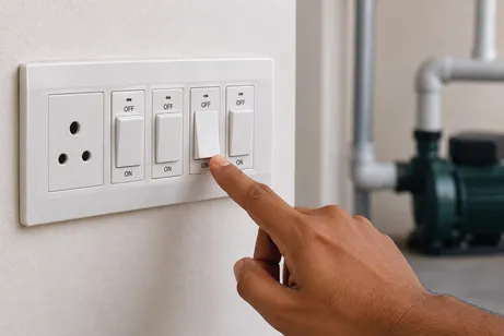 A finger pressing a switch on a wall switchboard, with a water pump behind it